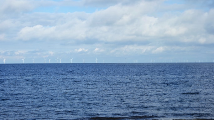 A tranquil sea with a wide expanse of blue water stretches towards the horizon, where numerous wind turbines stand against a backdrop of soft, cloudy skies. The scene conveys a sense of calm and harnesses the theme of renewable energy.