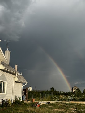 A rainbow arcs across a dark, cloudy sky above a garden area. A house on the left has a gray roof and white siding. In the foreground, there is a lawnmower and various garden tools. Beyond the fence, there are trees and another structure in the distance.
