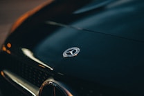 The image displays a close-up view of the emblem on the hood of a Mercedes-Benz car, with subtle reflections on the glossy, dark paint. The scene is slightly dimly lit, emphasizing the luxury and elegance associated with the vehicle.