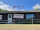 A rustic wooden building houses a laundromat with signs for wash, dry, and fold services. The signage prominently features red and blue text. Red cloth pieces are attached to antennas on the roof. The contrast between the blue sky, white clouds, and green foliage creates a quaint atmosphere.