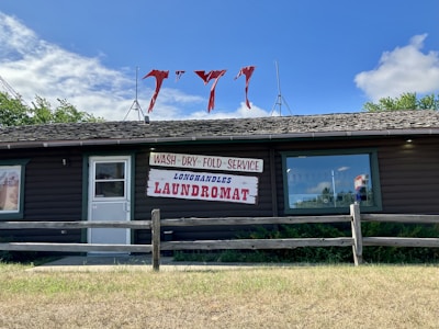 A rustic wooden building houses a laundromat with signs for wash, dry, and fold services. The signage prominently features red and blue text. Red cloth pieces are attached to antennas on the roof. The contrast between the blue sky, white clouds, and green foliage creates a quaint atmosphere.