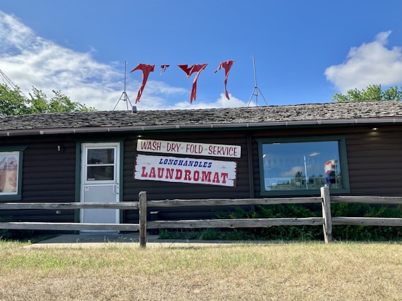 A rustic wooden building houses a laundromat with signs for wash, dry, and fold services. The signage prominently features red and blue text. Red cloth pieces are attached to antennas on the roof. The contrast between the blue sky, white clouds, and green foliage creates a quaint atmosphere.