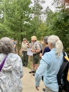 A group of people are gathered outdoors in a forested area, with tall trees in the background. One person, who appears to be a guide or ranger, is speaking into a microphone and holding some papers. The guide or ranger is dressed in a uniform and is wearing a backpack, while the listeners are in casual attire. The atmosphere seems to be educational and informative.