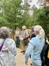 A group of people are gathered outdoors in a forested area, with tall trees in the background. One person, who appears to be a guide or ranger, is speaking into a microphone and holding some papers. The guide or ranger is dressed in a uniform and is wearing a backpack, while the listeners are in casual attire. The atmosphere seems to be educational and informative.