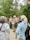 A group of people are gathered outdoors in a forested area, with tall trees in the background. One person, who appears to be a guide or ranger, is speaking into a microphone and holding some papers. The guide or ranger is dressed in a uniform and is wearing a backpack, while the listeners are in casual attire. The atmosphere seems to be educational and informative.