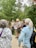 A group of people are gathered outdoors in a forested area, with tall trees in the background. One person, who appears to be a guide or ranger, is speaking into a microphone and holding some papers. The guide or ranger is dressed in a uniform and is wearing a backpack, while the listeners are in casual attire. The atmosphere seems to be educational and informative.