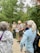 A group of people are gathered outdoors in a forested area, with tall trees in the background. One person, who appears to be a guide or ranger, is speaking into a microphone and holding some papers. The guide or ranger is dressed in a uniform and is wearing a backpack, while the listeners are in casual attire. The atmosphere seems to be educational and informative.