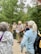 A group of people are gathered outdoors in a forested area, with tall trees in the background. One person, who appears to be a guide or ranger, is speaking into a microphone and holding some papers. The guide or ranger is dressed in a uniform and is wearing a backpack, while the listeners are in casual attire. The atmosphere seems to be educational and informative.