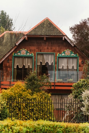 A charming, rustic house with a steeply pitched roof and textured shingle siding. The upper windows are adorned with floral decorations and lace curtains. Surrounding the house are lush green bushes and trees, and a black metal fence runs along the front.