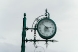 A vintage-style clock is mounted on an ornate metal post, featuring Roman numerals and decorative elements such as spirals and finials. The clock face displays two hands and a signature, suggesting a classic and elegant design.