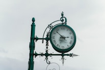 A vintage-style clock is mounted on an ornate metal post, featuring Roman numerals and decorative elements such as spirals and finials. The clock face displays two hands and a signature, suggesting a classic and elegant design.
