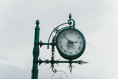 A vintage-style clock is mounted on an ornate metal post, featuring Roman numerals and decorative elements such as spirals and finials. The clock face displays two hands and a signature, suggesting a classic and elegant design.