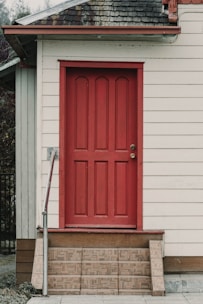A wooden building features a prominent red door, surrounded by cream-colored siding. The door is located at the top of a short set of patterned steps. The roof has a mixture of brown and gray shingles, and there is a metal handrail on the left side of the steps.