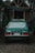 A green vintage Volvo car is parked on a gravel path in front of a rustic wooden cabin. The vehicle's license plate indicates it is from Chile. Surrounding the scene is abundant greenery with a variety of plants and trees.