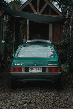 A green vintage Volvo car is parked on a gravel path in front of a rustic wooden cabin. The vehicle's license plate indicates it is from Chile. Surrounding the scene is abundant greenery with a variety of plants and trees.