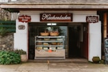 A quaint bakery storefront with a wooden sign reading 'Kuchenladen.' The entrance displays a glass case filled with various cakes and pastries. Above the case, wicker baskets add a rustic touch. The building exterior features stonework and plants, contributing to a cozy, traditional feel.