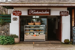 A quaint bakery storefront with a wooden sign reading 'Kuchenladen.' The entrance displays a glass case filled with various cakes and pastries. Above the case, wicker baskets add a rustic touch. The building exterior features stonework and plants, contributing to a cozy, traditional feel.