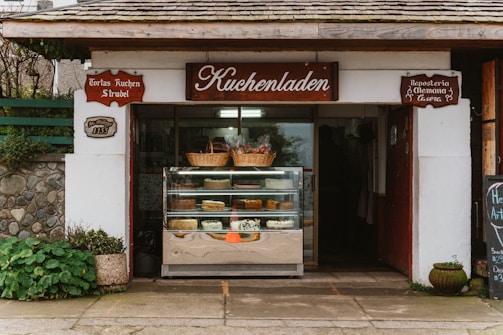 A quaint bakery storefront with a wooden sign reading 'Kuchenladen.' The entrance displays a glass case filled with various cakes and pastries. Above the case, wicker baskets add a rustic touch. The building exterior features stonework and plants, contributing to a cozy, traditional feel.
