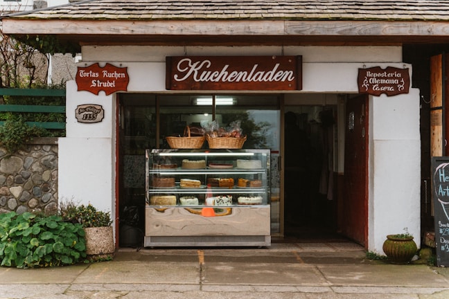A quaint bakery storefront with a wooden sign reading 'Kuchenladen.' The entrance displays a glass case filled with various cakes and pastries. Above the case, wicker baskets add a rustic touch. The building exterior features stonework and plants, contributing to a cozy, traditional feel.