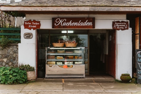 A quaint bakery storefront with a wooden sign reading 'Kuchenladen.' The entrance displays a glass case filled with various cakes and pastries. Above the case, wicker baskets add a rustic touch. The building exterior features stonework and plants, contributing to a cozy, traditional feel.