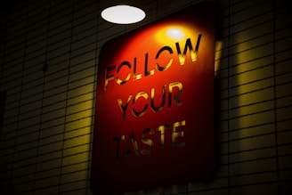 A red rectangular sign with the words 'FOLLOW YOUR TASTE' cut out, illuminated by a light from above. The sign is mounted on a wall with a pattern of horizontal lines, creating a contrast between the light and shadows.