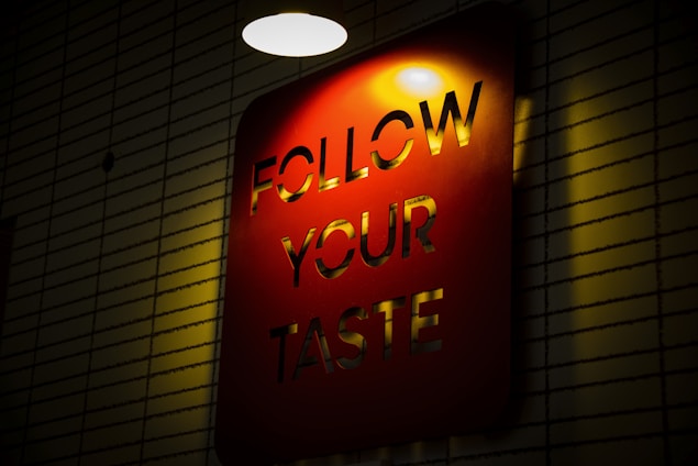 A red rectangular sign with the words 'FOLLOW YOUR TASTE' cut out, illuminated by a light from above. The sign is mounted on a wall with a pattern of horizontal lines, creating a contrast between the light and shadows.