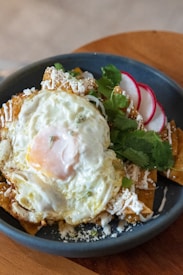 A plate of Mexican-style chilaquiles topped with a fried egg, garnished with slices of radish, fresh cilantro, and crumbled cheese. The dish is served in a dark, round plate set on a wooden surface.