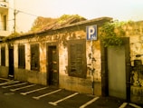 An old, weathered building with peeling paint and dark shutters on its windows. The structure is overgrown with greenery on the roof, indicating age and neglect. There is a parking sign indicating a bicycle parking area in front of the building. The overall setting suggests an urban environment with limited maintenance.