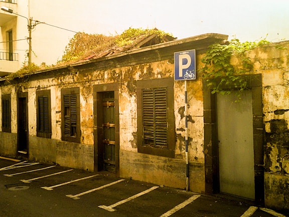 An old, weathered building with peeling paint and dark shutters on its windows. The structure is overgrown with greenery on the roof, indicating age and neglect. There is a parking sign indicating a bicycle parking area in front of the building. The overall setting suggests an urban environment with limited maintenance.