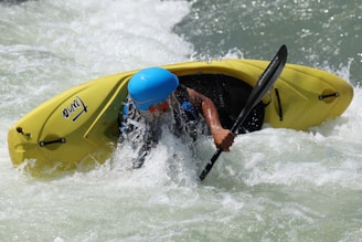 A person wearing a blue helmet is engaged in kayaking on a river with whitewater rapids. The individual is navigating the turbulent waters with a black paddle, while the yellow kayak rides the waves.