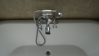 A chrome faucet is mounted on a tiled wall above a white bathtub. The faucet includes hot and cold knobs, a spout, and an attached shower head with a flexible metal hose. The wall appears to have a beige, textured surface.