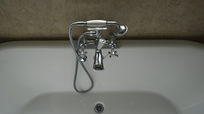 A chrome faucet is mounted on a tiled wall above a white bathtub. The faucet includes hot and cold knobs, a spout, and an attached shower head with a flexible metal hose. The wall appears to have a beige, textured surface.