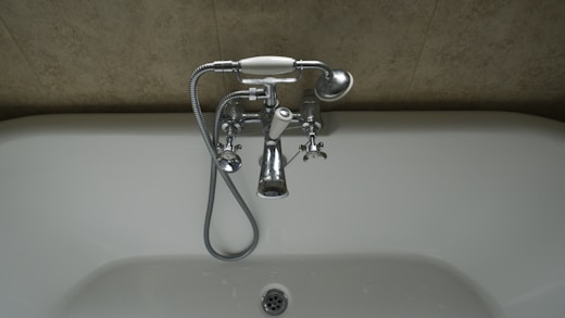 A chrome faucet is mounted on a tiled wall above a white bathtub. The faucet includes hot and cold knobs, a spout, and an attached shower head with a flexible metal hose. The wall appears to have a beige, textured surface.