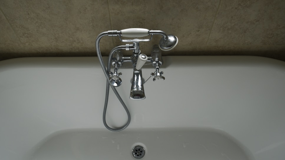 A chrome faucet is mounted on a tiled wall above a white bathtub. The faucet includes hot and cold knobs, a spout, and an attached shower head with a flexible metal hose. The wall appears to have a beige, textured surface.