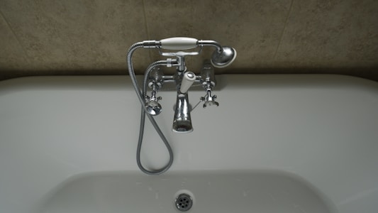 A chrome faucet is mounted on a tiled wall above a white bathtub. The faucet includes hot and cold knobs, a spout, and an attached shower head with a flexible metal hose. The wall appears to have a beige, textured surface.