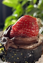 A close-up of a chocolate dessert topped with a large, ripe strawberry. The chocolate appears rich and glossy, with specks of green pistachios embedded in it. The background is blurred, emphasizing the dessert's texture and vivid colors.