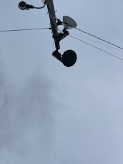 A utility pole seen from below, with multiple devices attached including a surveillance camera, a streetlight, a satellite dish, and a round-shaped device. Several wires are extending from the pole into different directions. The sky in the background is overcast with grey clouds.