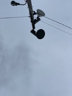 A utility pole seen from below, with multiple devices attached including a surveillance camera, a streetlight, a satellite dish, and a round-shaped device. Several wires are extending from the pole into different directions. The sky in the background is overcast with grey clouds.