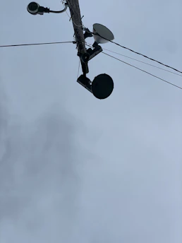 A utility pole seen from below, with multiple devices attached including a surveillance camera, a streetlight, a satellite dish, and a round-shaped device. Several wires are extending from the pole into different directions. The sky in the background is overcast with grey clouds.
