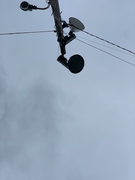 A utility pole seen from below, with multiple devices attached including a surveillance camera, a streetlight, a satellite dish, and a round-shaped device. Several wires are extending from the pole into different directions. The sky in the background is overcast with grey clouds.