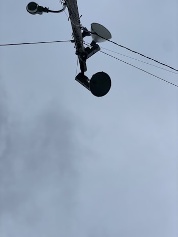 A utility pole seen from below, with multiple devices attached including a surveillance camera, a streetlight, a satellite dish, and a round-shaped device. Several wires are extending from the pole into different directions. The sky in the background is overcast with grey clouds.