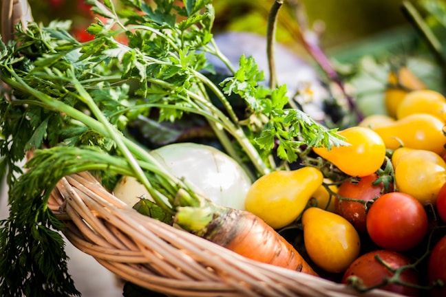 A wicker basket is filled with an assortment of fresh vegetables, including yellow pear-shaped tomatoes, red cherry tomatoes, a round white onion, and leafy sprigs of green herbs. The sunlight creates a vibrant display of colors, highlighting the freshness of the produce.
