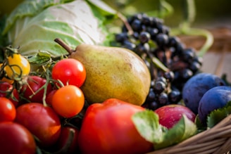 A variety of fresh fruits and vegetables including tomatoes, a pear, grapes, and plums are arranged in a basket. The scene is colorful and vibrant, with the focus on the natural textures and colors of the produce.