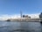 A waterfront view features a tall skyscraper and multiple modern commercial buildings under a clear blue sky. The water in the foreground is calm, reflecting some of the sky's hues. A dock extends along the waterfront, possibly serving as a terminal or a pier.