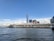 A waterfront view features a tall skyscraper and multiple modern commercial buildings under a clear blue sky. The water in the foreground is calm, reflecting some of the sky's hues. A dock extends along the waterfront, possibly serving as a terminal or a pier.