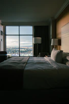 Cozy hotel room with warm lighting and a comfortable bed near a window showing a city view at sunset.