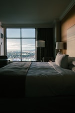 A dimly lit hotel room features a neatly made bed with white sheets and a dark blanket. Two bedside lamps flank the bed, casting a soft glow. A large window offers a view of a cityscape during twilight, with the lights from buildings and streets visible in the distance.