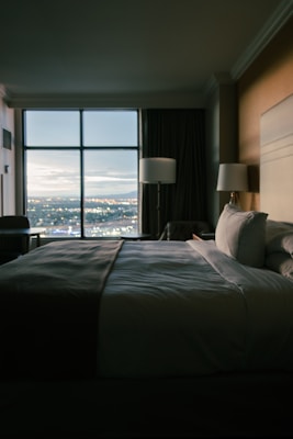A cozy hotel room with a large bed, soft lighting, and a window showing a cityscape at dusk.
