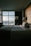 A dimly lit hotel room features a neatly made bed with white sheets and a dark blanket. Two bedside lamps flank the bed, casting a soft glow. A large window offers a view of a cityscape during twilight, with the lights from buildings and streets visible in the distance.