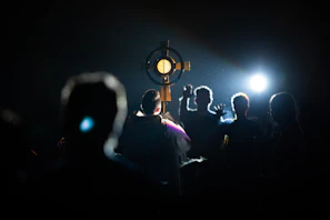 A silhouette of a person holding a large, circular religious object, possibly a monstrance, with a lit candle or light source at its center. Surrounding the central figure are several other silhouetted individuals, with the scene illuminated from behind by a bright light, creating a dramatic and spiritual atmosphere.